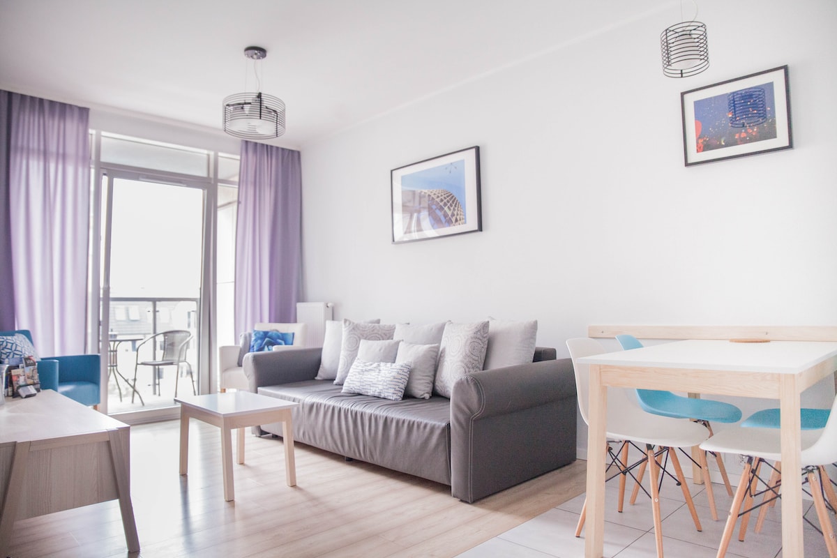 A bright living area features a comfortable sofa adorned with cushions, accompanied by a wooden coffee table. A dining table with colorful chairs sits nearby. Natural light enters through large glass doors, framed by soft curtains, leading to a private balcony.