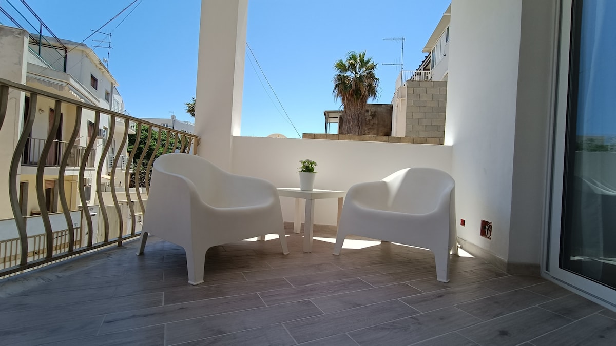 A private balcony is presented with two white chairs and a small table, positioned under clear blue skies. A potted plant adds a touch of greenery, while a palm tree and the neighboring buildings can be seen in the background, enhancing the inviting outdoor space.