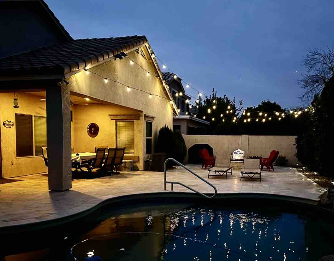 An outdoor entertaining space is depicted in the evening, featuring a pool with a gentle glow. String lights are draped overhead, illuminating a dining table surrounded by chairs and a fire pit. Two red lounge chairs are placed invitingly by the pool's edge.