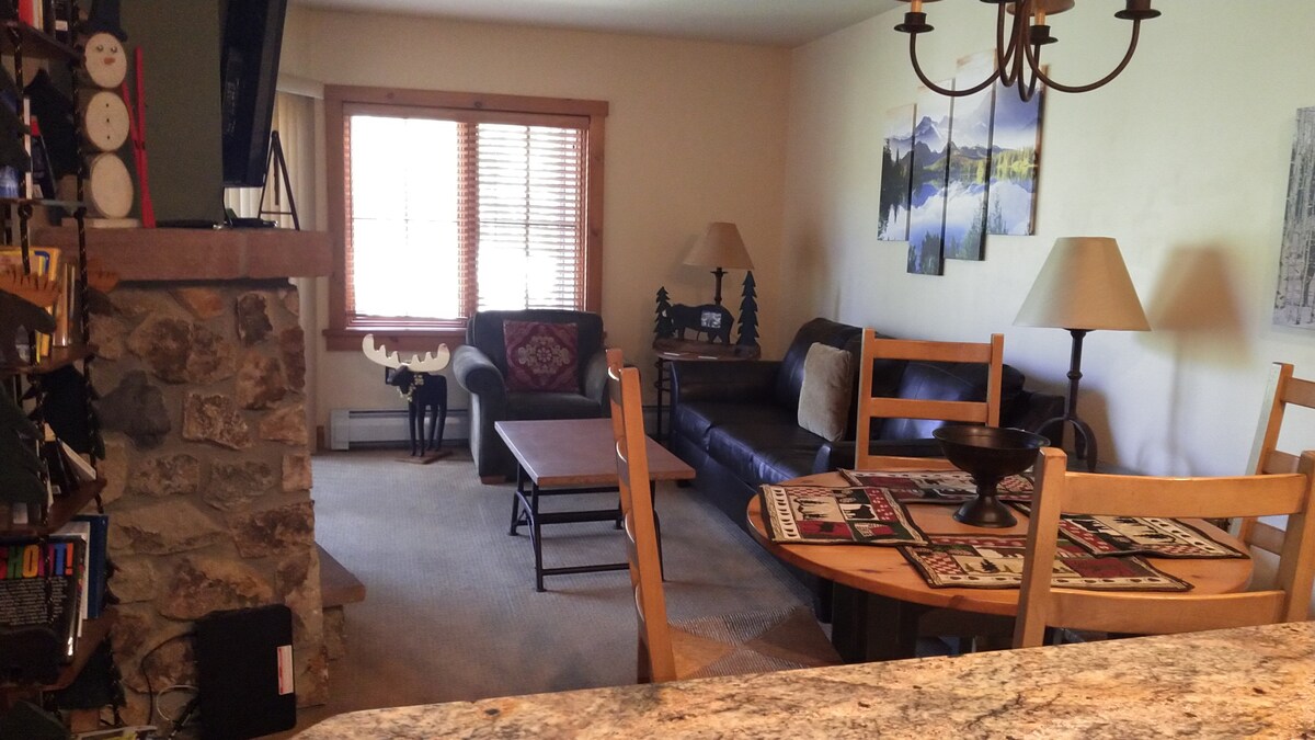 The living area showcases a comfortable seating arrangement, including a dark leather sofa, a coffee table, and two lamps providing warm light. A wooden dining table with four chairs is nearby. Scenic mountain wall art and large windows allow natural light into the space.