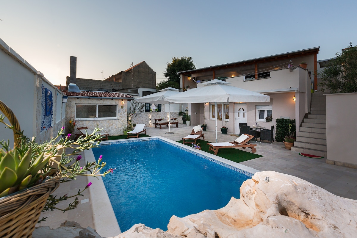 An outdoor swimming pool is centrally located, surrounded by sun loungers. A patio area is visible, featuring a seating arrangement under a large umbrella. The modern architecture includes a two-story building with multiple windows, providing an open and welcoming atmosphere.