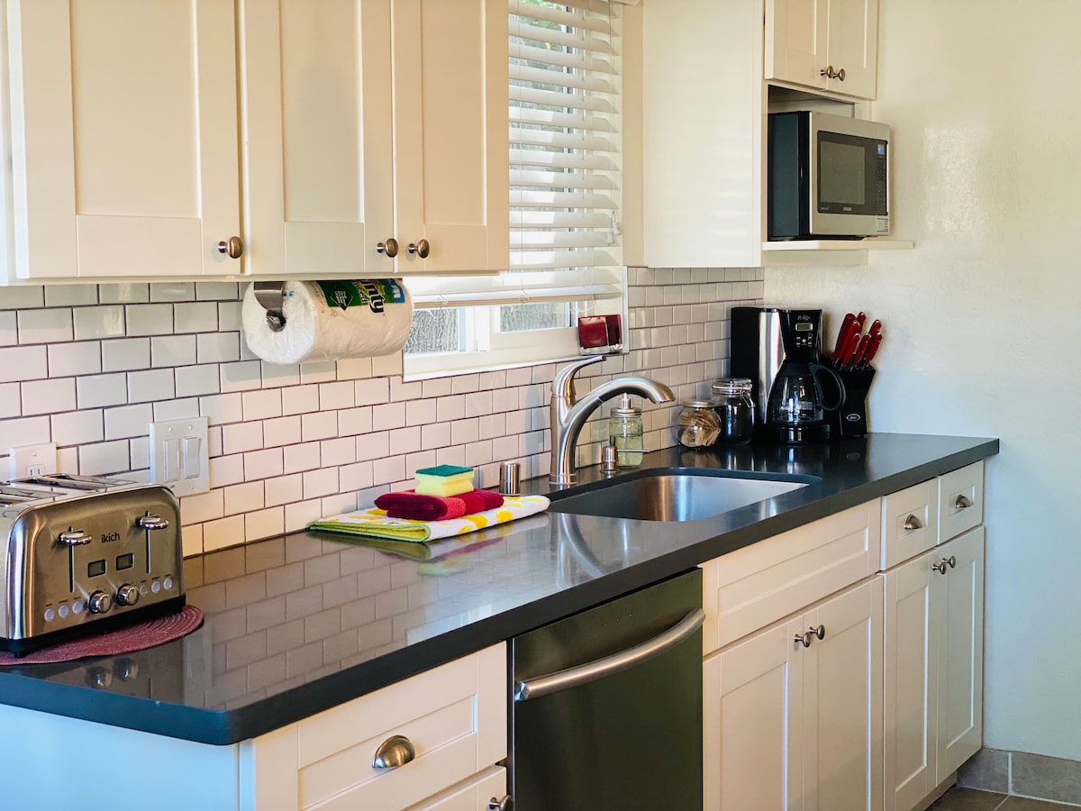 The kitchen features white cabinetry and a sleek countertop with a stainless steel sink. Appliances include a microwave and coffee maker, alongside a toaster. Natural light floods in through a window, while colorful dish towels add a touch of vibrancy.