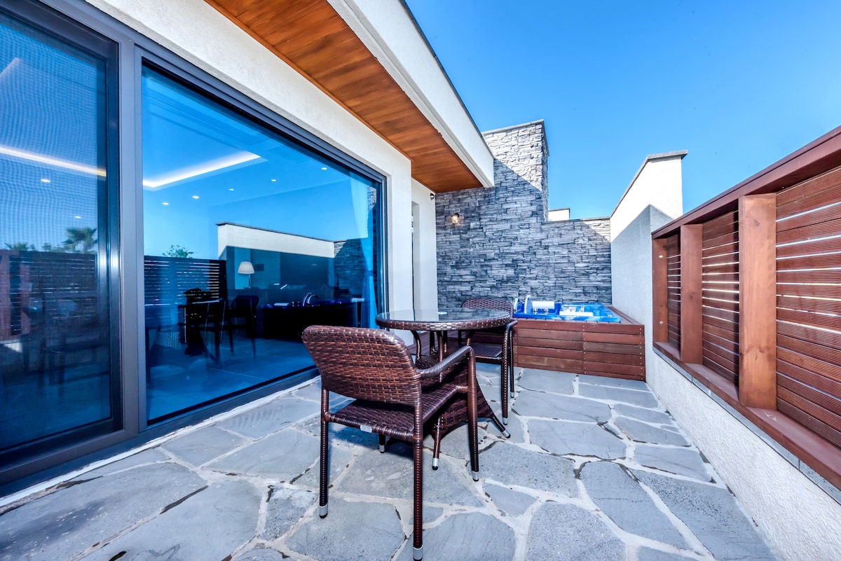 A private terrace features outdoor seating with two wicker chairs set on a stone floor. The terrace overlooks a compact outdoor spa, partially enclosed by a stone wall, with large glass doors providing access to the living space inside.