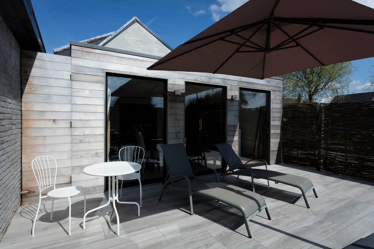 A private terrace is shown, featuring two lounge chairs positioned beside a small round table. A large umbrella provides shade, and the wooden paneling of the suite reflects a modern design. Floor tiles enhance the outdoor space, inviting relaxation.