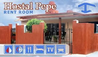The exterior of a red and beige building is showcased, featuring a gated entrance and vibrant plant life. Icon signs indicate available amenities such as water, air conditioning, dining, television, and garage access. The structure is complemented by a clear blue sky.