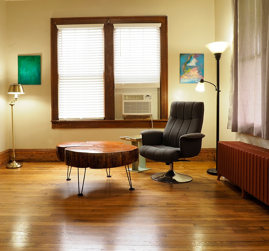 A spacious seating area featuring a unique wooden coffee table complemented by a modern upholstered chair. Natural light filters through two windows with blinds. Artwork adorns the walls, and a floor lamp provides additional lighting. An air conditioning unit is visible in the window.