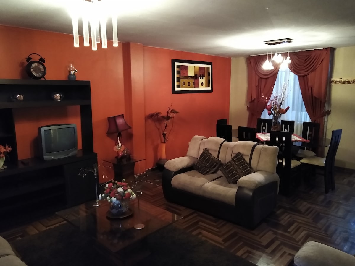 A cozy living space features a mix of neutral and vibrant orange walls. A comfortable seating area includes a light-colored sofa set arranged around a wooden coffee table. A dining area with a dark wood table and six chairs is visible, complemented by decorative plants and wall art.