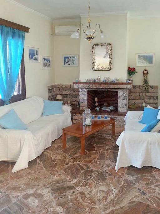 It is a traditional Greek style living room with a fireplace that adds to the feeling of coziness. There is a mirror wall on top of the fireplace and the light of the sun enters through big windows.