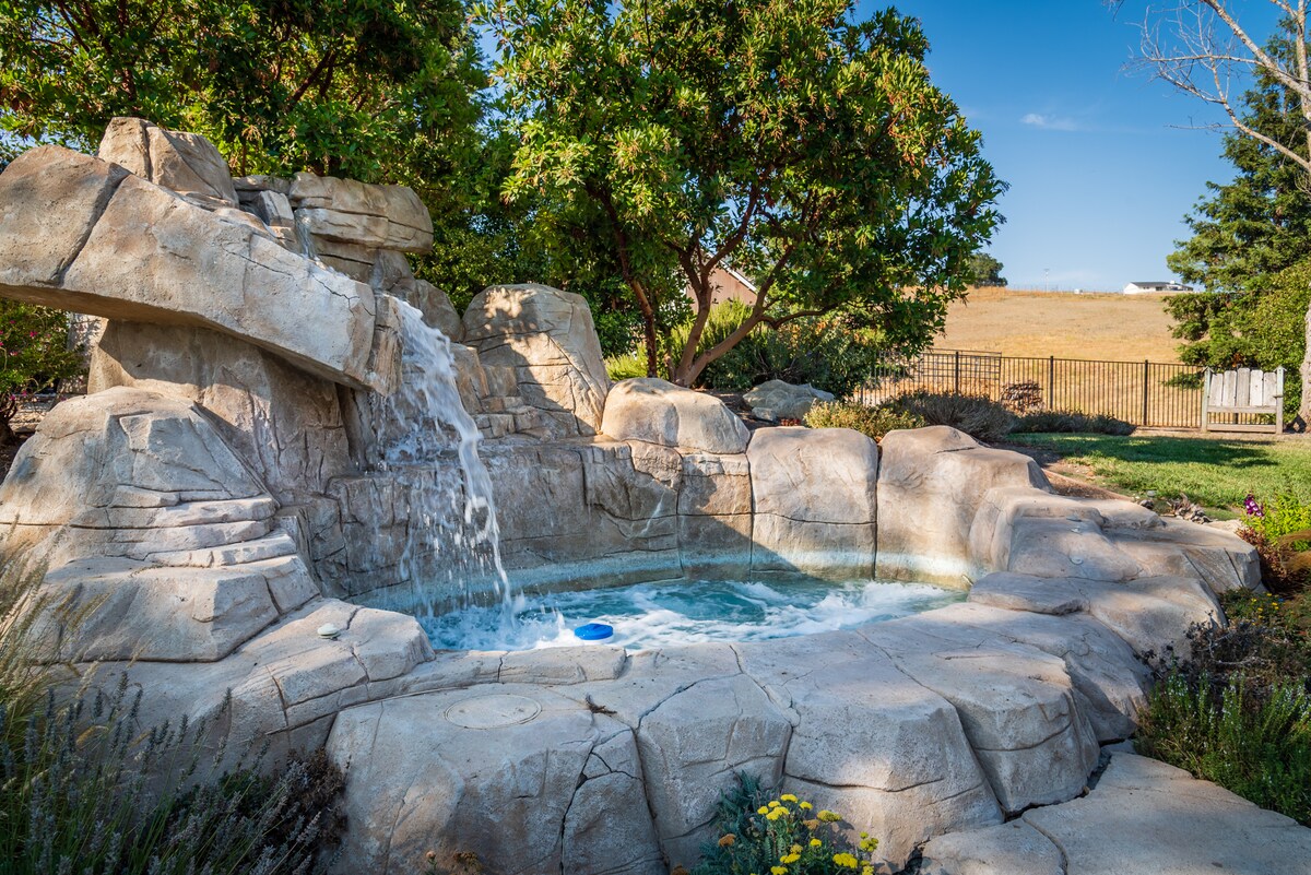 A natural stone spa is set amidst lush greenery, featuring a gentle waterfall that cascades into the water. The surrounding area includes bushes and flowers, providing a serene atmosphere for relaxation.