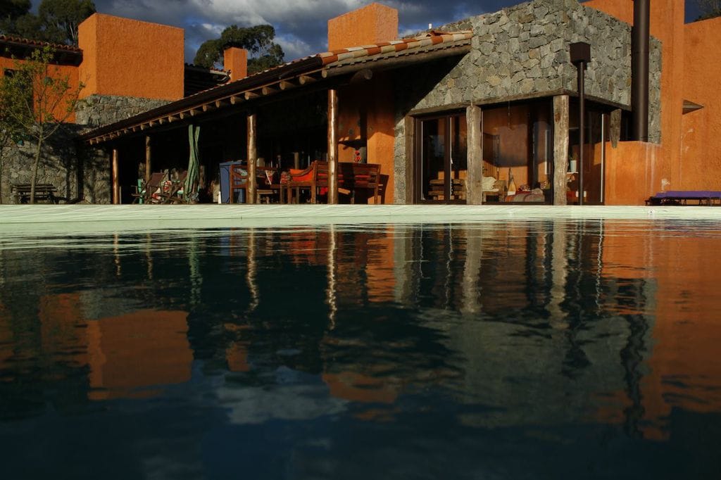 The house's exterior is visible, featuring warm orange walls and rustic stone accents. A spacious terrace with wooden furniture is presented under a patio roof, while reflections in the tranquil pool enhance the serene ambiance. Lush greenery and mountains are subtly hinted in the background.