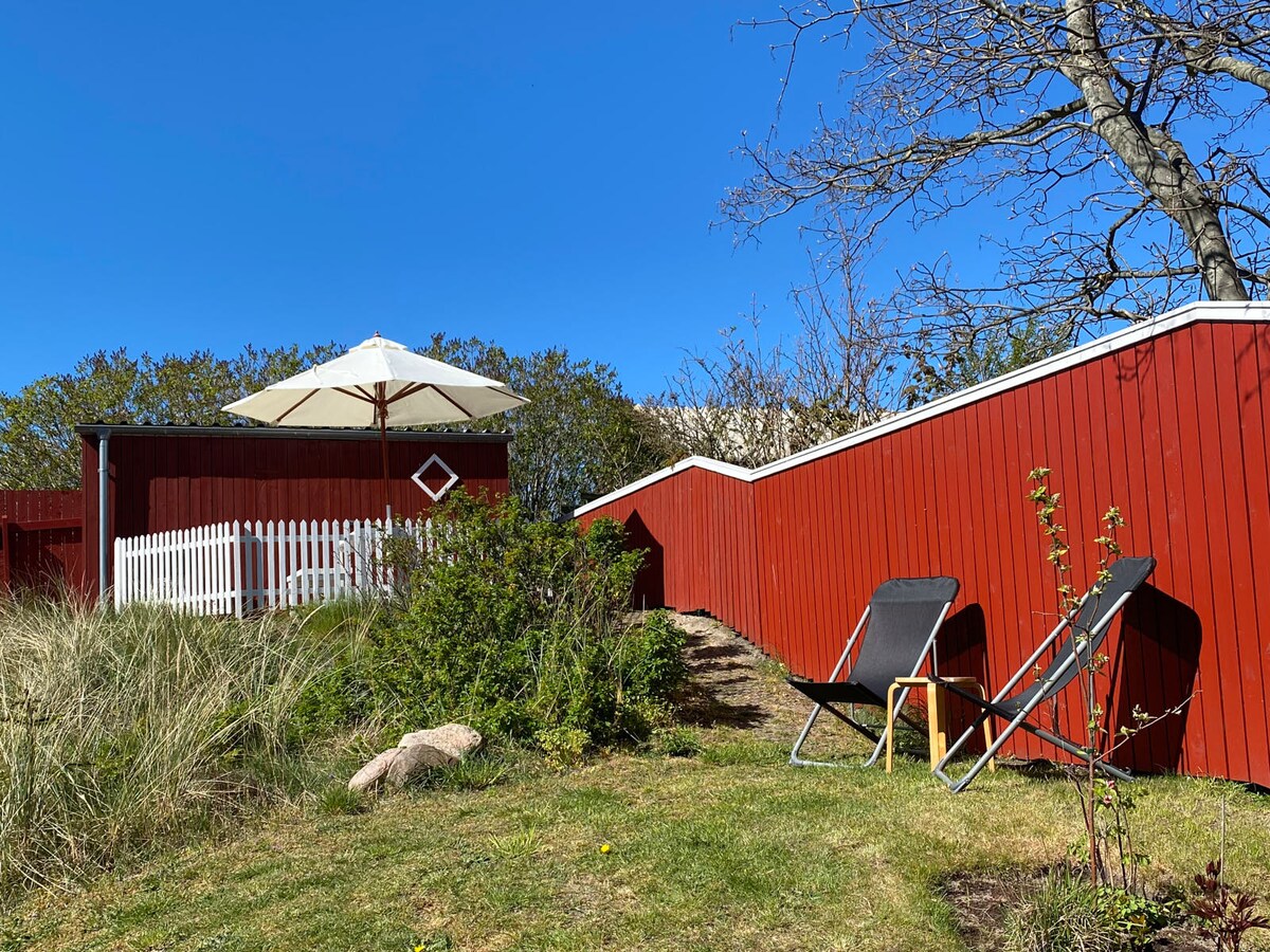 A private outdoor space is showcased with green grass and wooden decking. A large umbrella provides shade over a seating area. Two lounge chairs are positioned beside a small garden, surrounded by lush vegetation and a red wooden fence.