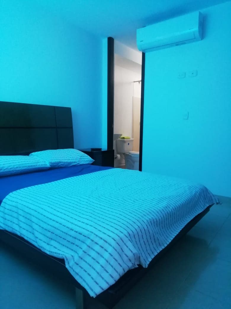 A bright bedroom features a spacious bed with a striped blanket. Soft blue lighting casts a calming ambiance. A doorway leads to a bathroom, while a small nightstand is positioned beside the bed. The walls and floor are light-colored, enhancing the room's openness.