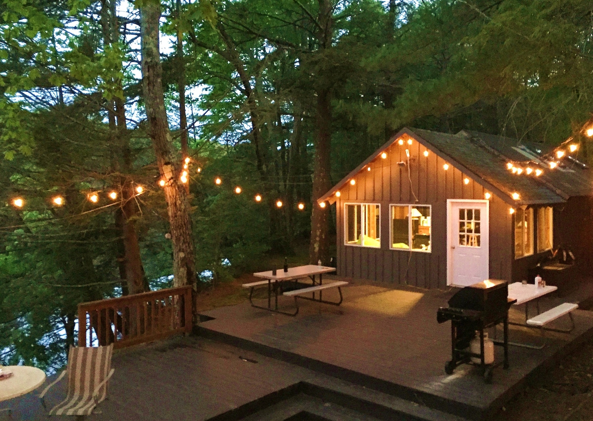 A cozy cabin sits nestled among trees, illuminated by warm string lights. A spacious deck features picnic tables and a propane grill, offering areas for outdoor dining and relaxation, with the tranquil water visible nearby.