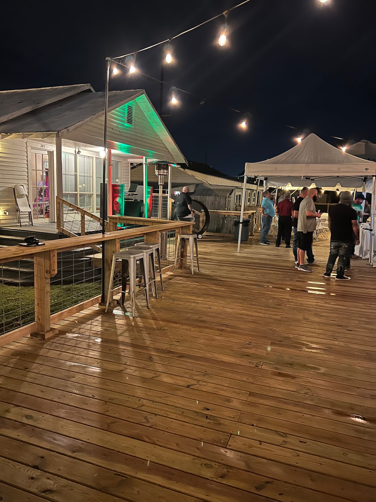 A well-lit deck area is shown at night, featuring wooden flooring and string lights overhead. Tents are set up for events, with people gathered and socializing. The cottage is visible in the background, reflecting a welcoming ambiance amidst the evening setting.