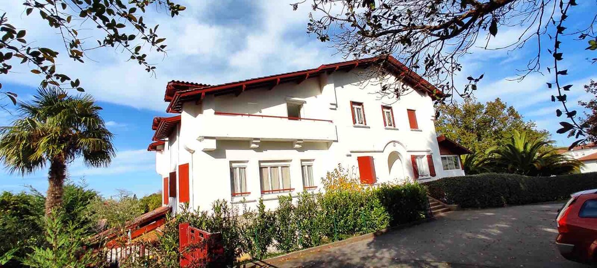 The apartment building is set within a landscaped garden featuring palm trees and hedges. A staircase leads to the entrance, framed by vibrant green flora. The light-colored exterior is contrasted by red accents, and the sky above is partly cloudy, showcasing a serene atmosphere.