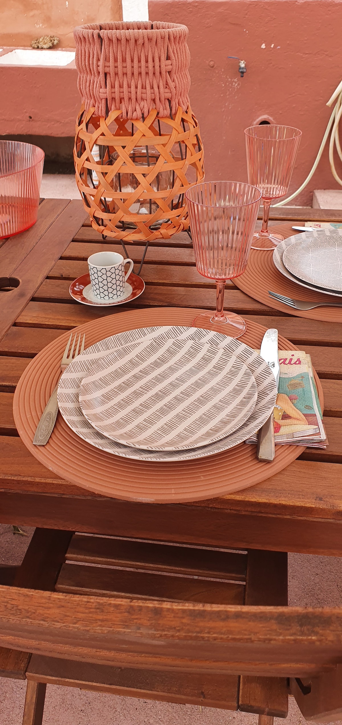 A wooden dining table is set with a place setting that includes patterned plates, silverware, and colorful glasses. An artistic lantern adds texture to the scene. A magazine is casually placed beside the plates, suggesting a relaxed dining atmosphere.
