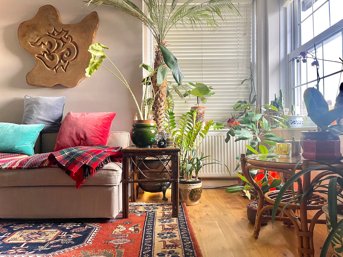 A cozy living space features a light-toned sofa adorned with colorful cushions, alongside a variety of plants, including palm and leafy greenery. A woven rug adds texture, while sunlight filters through the window, casting pleasant shadows on the wooden floor.