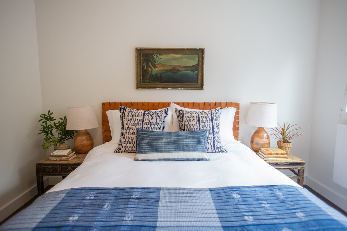 A cozy bedroom features a spacious bed with a variety of textured pillows, complemented by a decorative blue throw. Two bedside tables, each with a lamp, are positioned on either side. A piece of landscape art hangs above the bed, and a small plant adds a touch of greenery.
