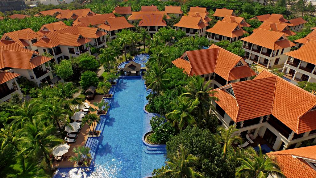 An aerial view captures the layout of the resort, featuring a central, blue swimming pool surrounded by lush greenery and palm trees. A series of villas with distinctive orange roofs are interspersed throughout the landscape, offering an inviting tropical atmosphere.