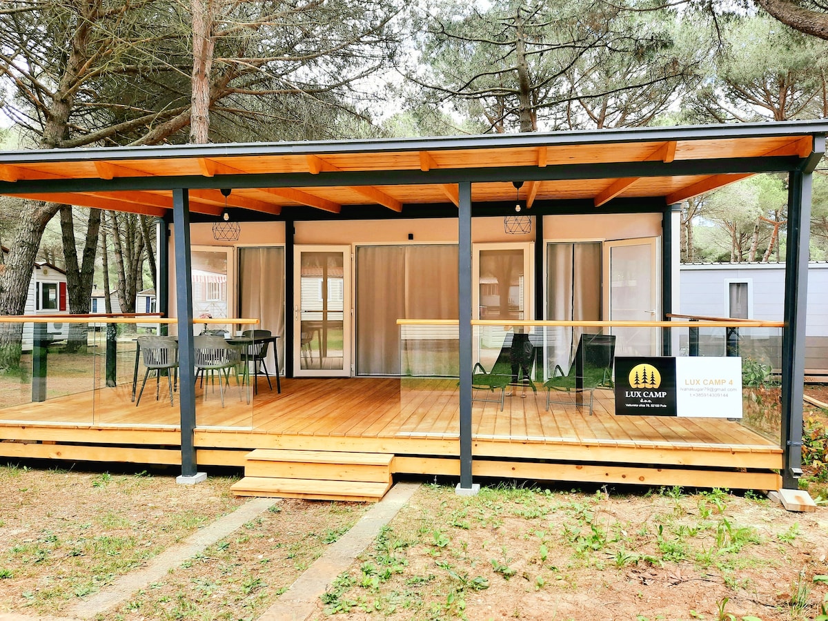 A wooden deck with a covered seating area is visible, featuring black chairs and a glass railing. The structure has large windows that provide natural light, and trees are present in the background, creating a serene outdoor setting.