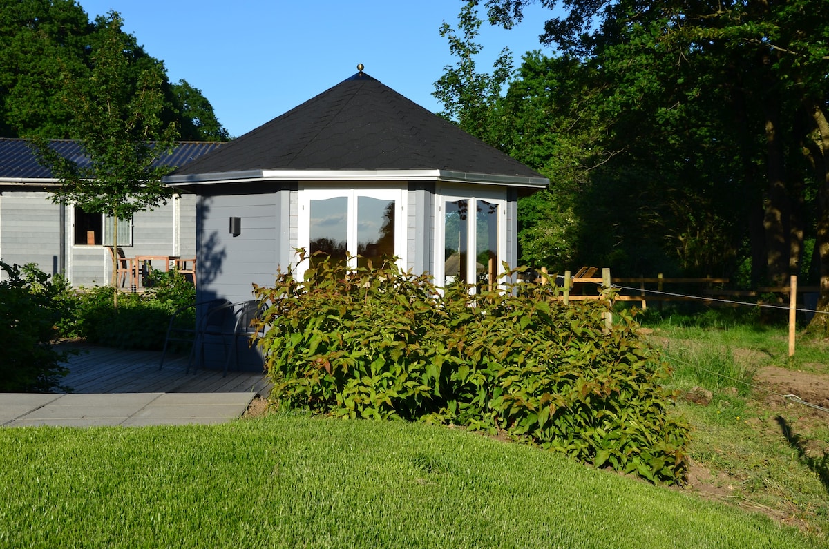 A contemporary cabin with a unique round shape is surrounded by lush greenery and a well-maintained lawn. Large windows offer views of the landscape, while a pathway leads to the inviting entrance. A nearby fence enhances the serene outdoor setting.
