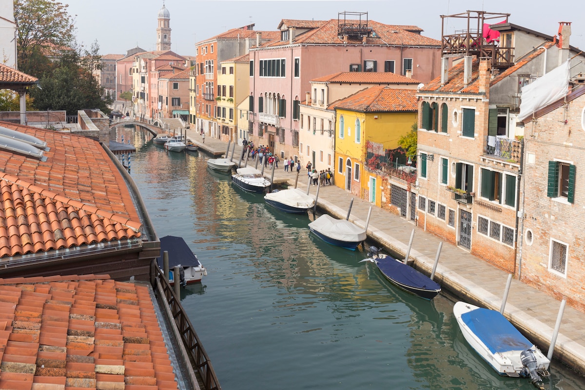 A serene canal is lined with colorful buildings and various boats, reflecting the charm of a typical Venetian neighborhood. Traditional architecture is visible, with terracotta roofs and vibrant facades. The tranquil water surface creates a calm atmosphere, inviting exploration of the area's rich culture.