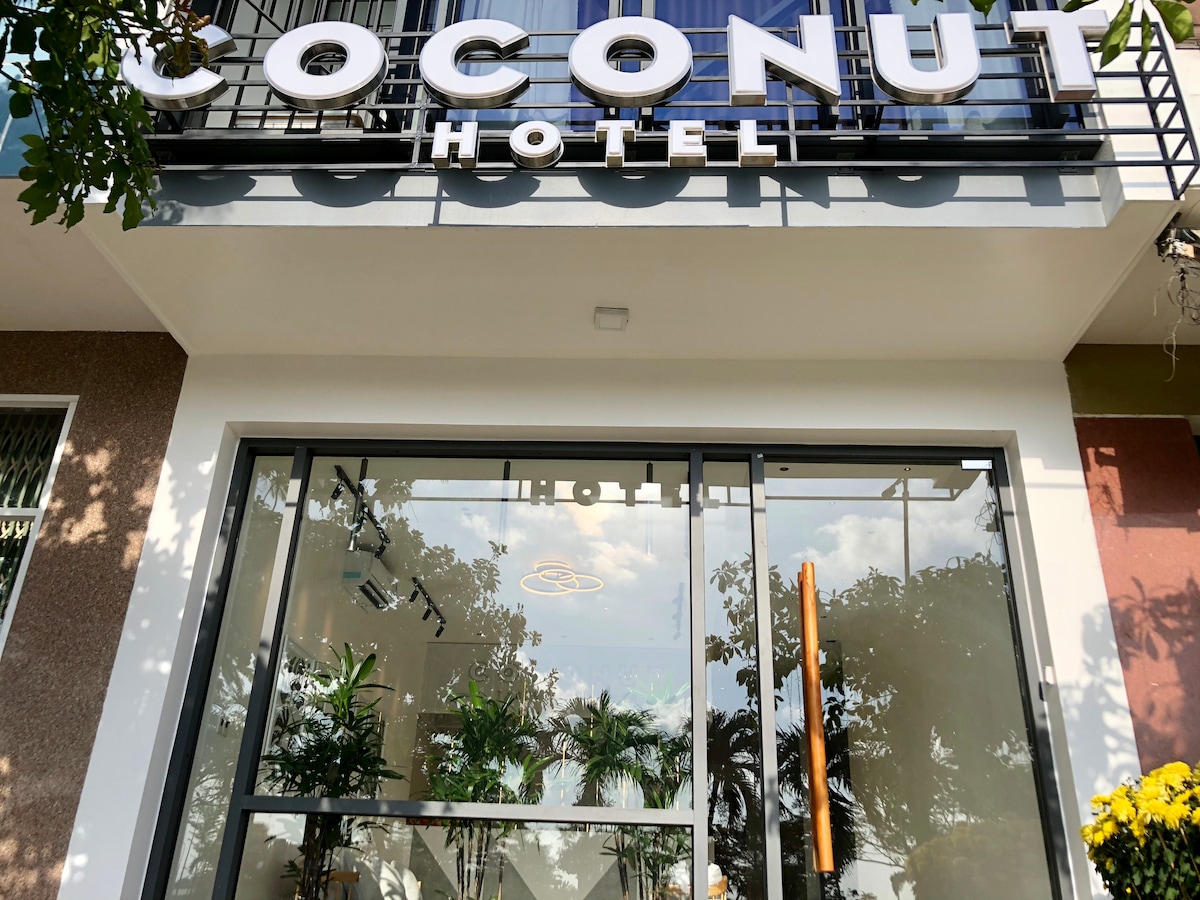 The hotel entrance features large glass doors with a modern frame, reflecting greenery from outside. The name 'Coconut Hotel' is prominently displayed above in bold letters. Potted plants are positioned near the entrance, contributing to a welcoming ambiance.