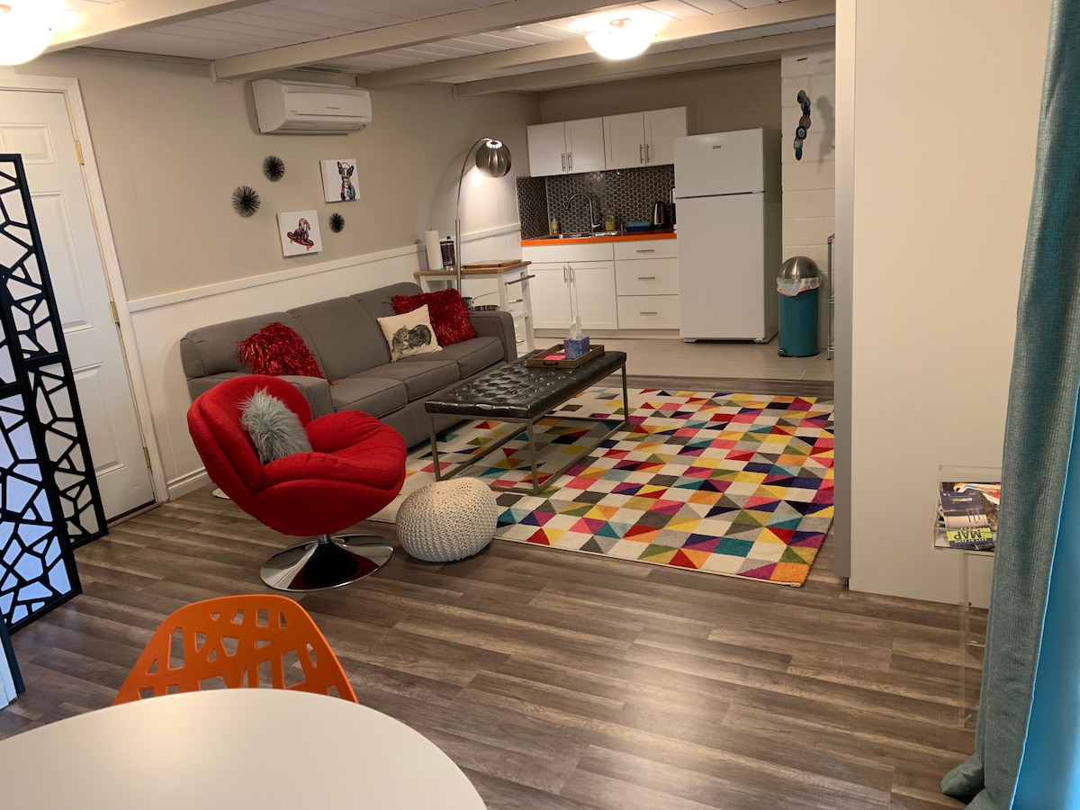 A cozy living space presents a gray sofa and a vibrant red chair, complemented by a multicolored area rug. An adjacent kitchenette is equipped with essential appliances, including a refrigerator and stove. The room is illuminated by overhead lighting, and wooden flooring adds warmth to the setting.