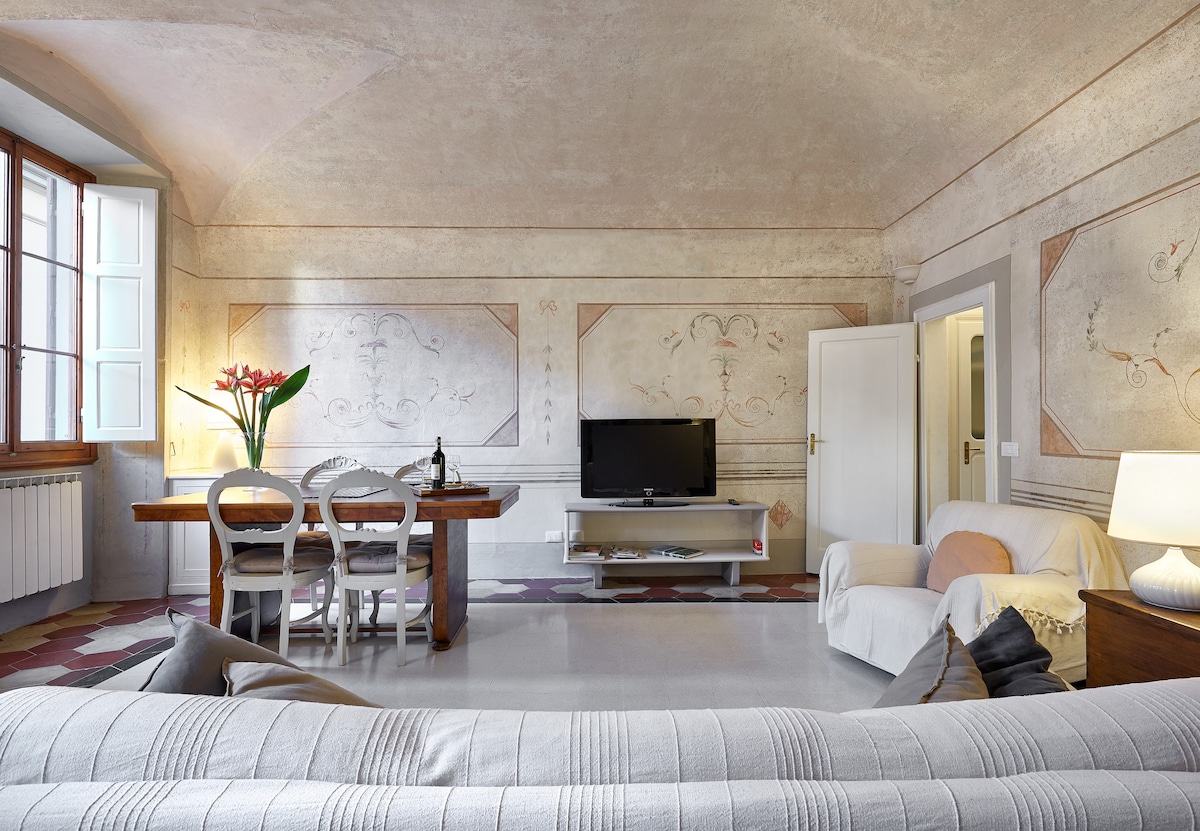 A spacious living area features a large window with light filtering through, illuminating the delicate wall frescoes. A dining table with chairs is set nearby, while a television rests on a low stand across from a comfortable sofa adorned with cushions.
