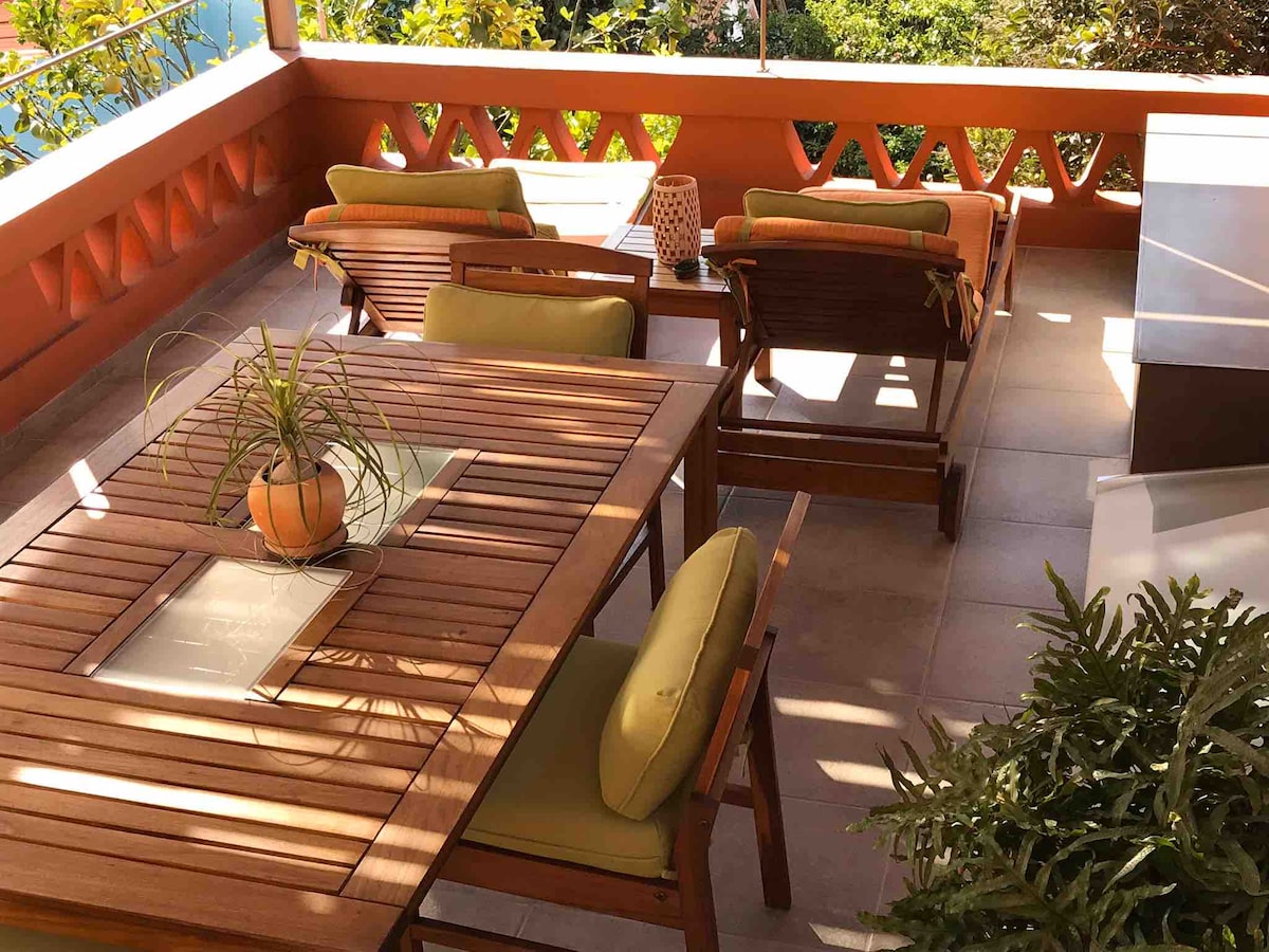 A covered terrace featuring a wooden table surrounded by four chairs, complemented by two deckchairs. A potted plant sits on the table, while natural light filters through the surrounding greenery creating a tranquil setting.