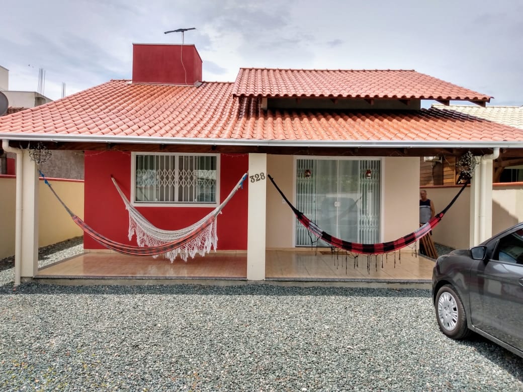 The exterior of the house features a terracotta roof with a welcoming front porch. Two hammocks are suspended between the columns, providing a relaxing spot. A gravel pathway leads to the entrance, with a parked car visible nearby. The overall structure presents a blend of red and neutral tones.