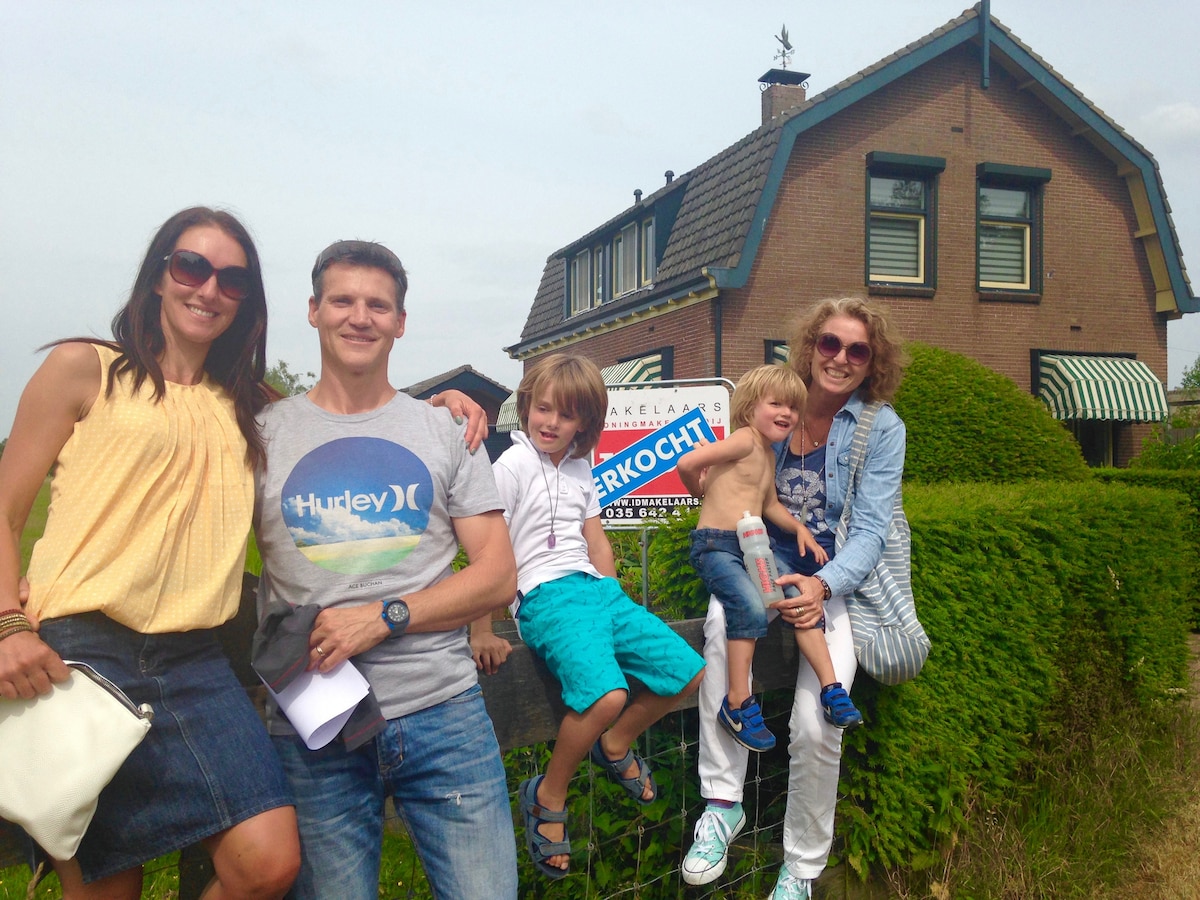A family poses outside a two-story home, with a green hedge in the foreground and a real estate sign in view. The house features a pitched roof and multiple windows with striped awnings. Guests are seated on a wooden bench, displaying a casual and friendly atmosphere.