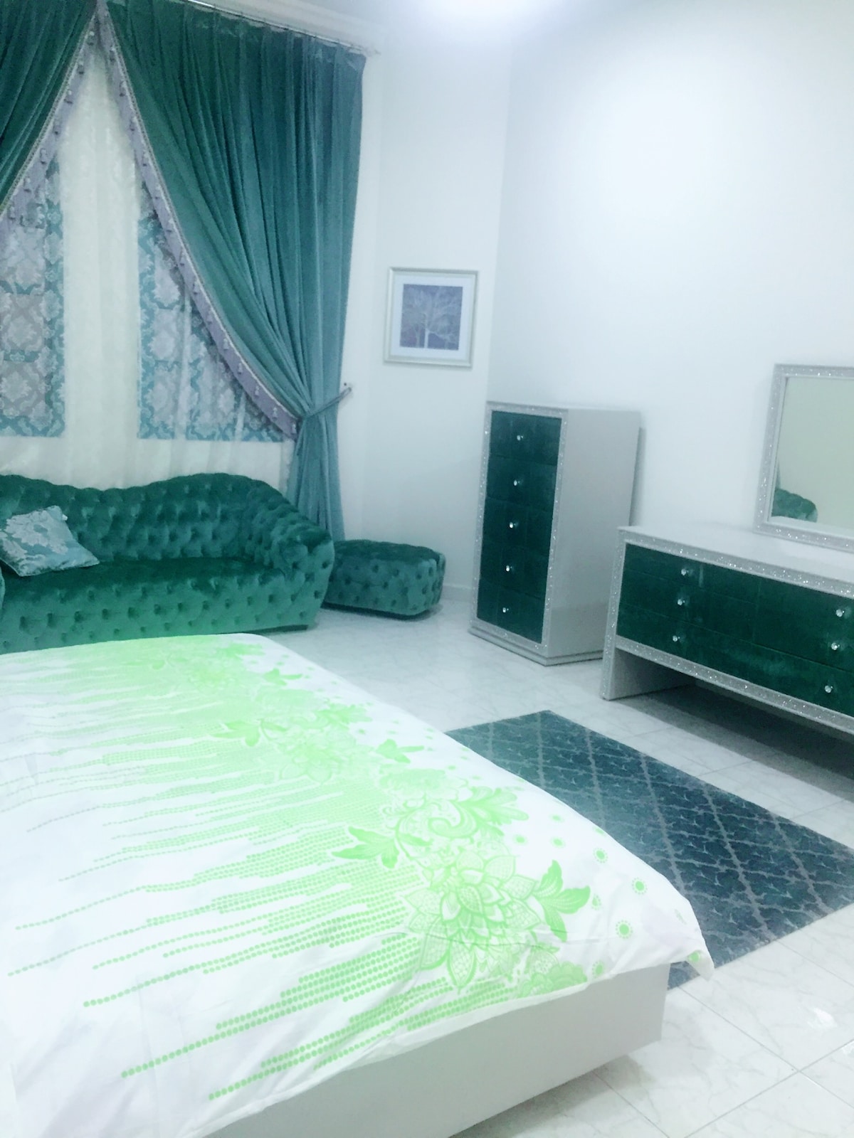 A bedroom featuring a spacious bed adorned with a green floral bedspread. A tufted green sofa and a matching armchair complement the decor. A mirrored dresser stands opposite the bed, while patterned curtains frame a window, allowing natural light to enter the room.