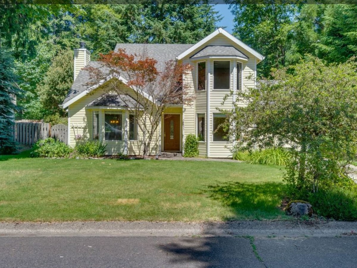 The cottage-style home is surrounded by lush greenery, featuring a well-maintained front yard. Large windows illuminate the inviting entrance, and a distinctive architecture showcases a mix of traditional and modern elements. The exterior is accented by a variety of trees and shrubs.
