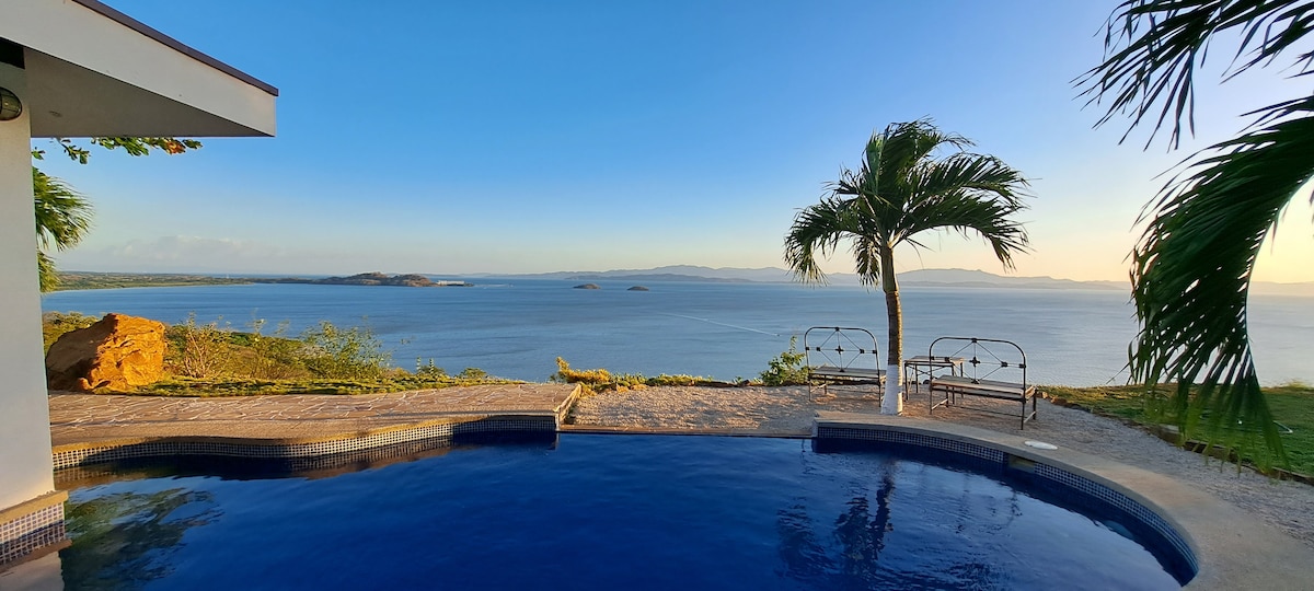An infinity pool is seen overlooking the ocean, framed by a lush palm tree. The calming blue waters of the Gulf of Nicoya stretch into the distance, with scattered islands visible on the horizon. A small terrace with seating areas complements the serene atmosphere.
