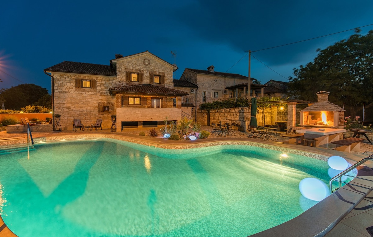 An illuminated shared pool features gentle curves, surrounded by lush greenery and a stone barbecue area. A historic building with a textured facade stands in the background, while patio seating offers spaces for relaxation. Soft blue lighting highlights the water, creating a calm ambiance.