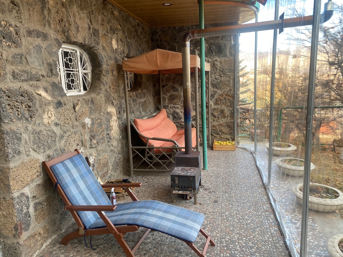 A screened porch area features a comfortable sofa with an orange cover and a relaxing lounge chair with a blue checkered pattern. A small wood-burning stove is positioned between them, and light enters through the glass panels, highlighting the stone walls.