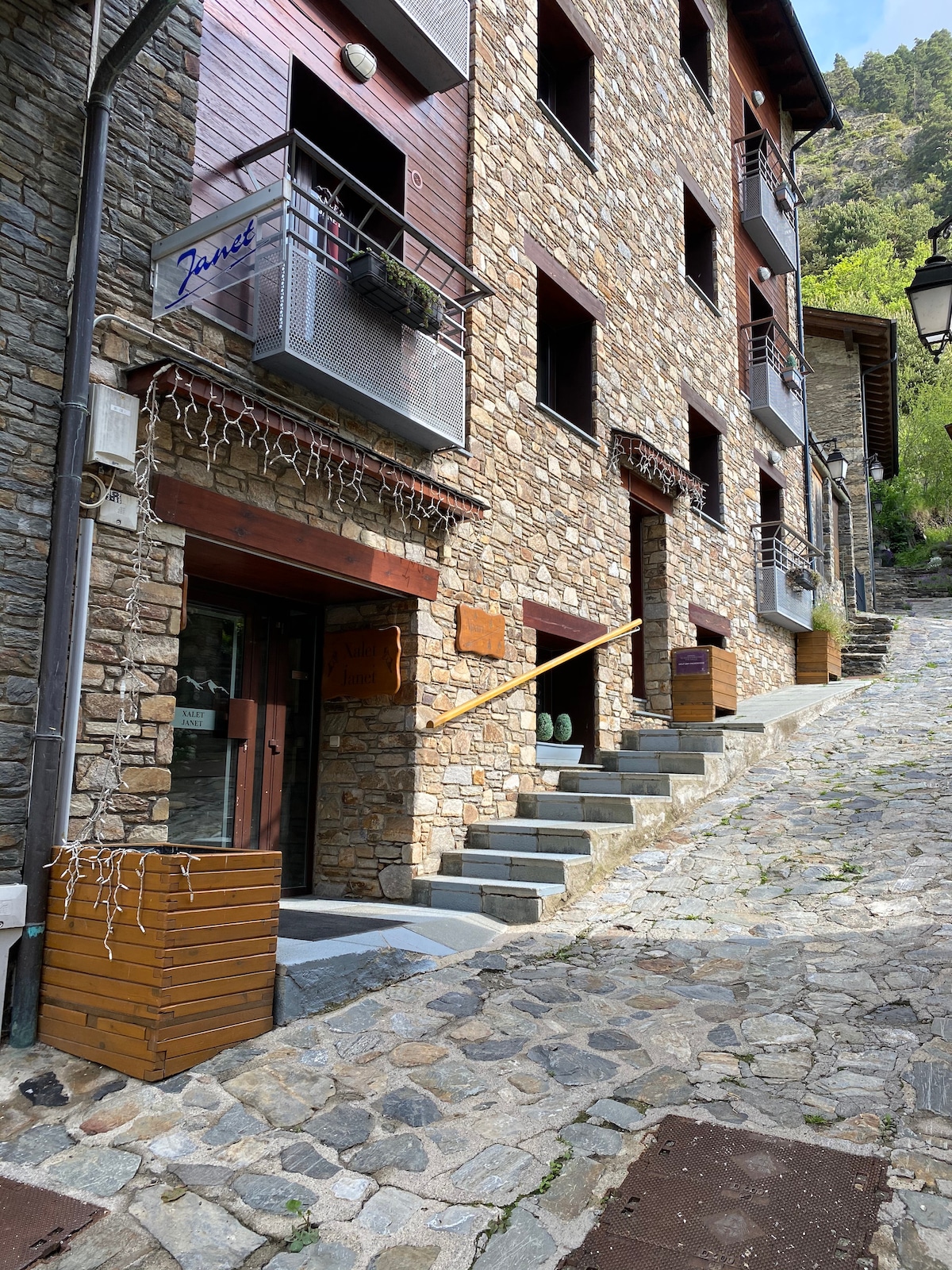 A charming stone building with multiple stories is seen along a cobblestone path. Balconies with planters adorn the upper levels, while the entrance features wooden signage. The natural surroundings add to the building's rustic appeal, highlighting a welcoming environment.