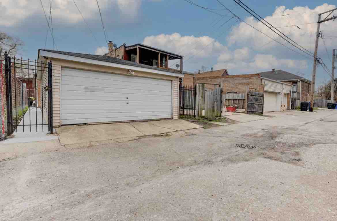 The image displays a garage with a white door, situated on a gravel alley. In the background, buildings can be seen, along with a wooden fence and power lines overhead. A clear sky with a few clouds creates a calm backdrop.
