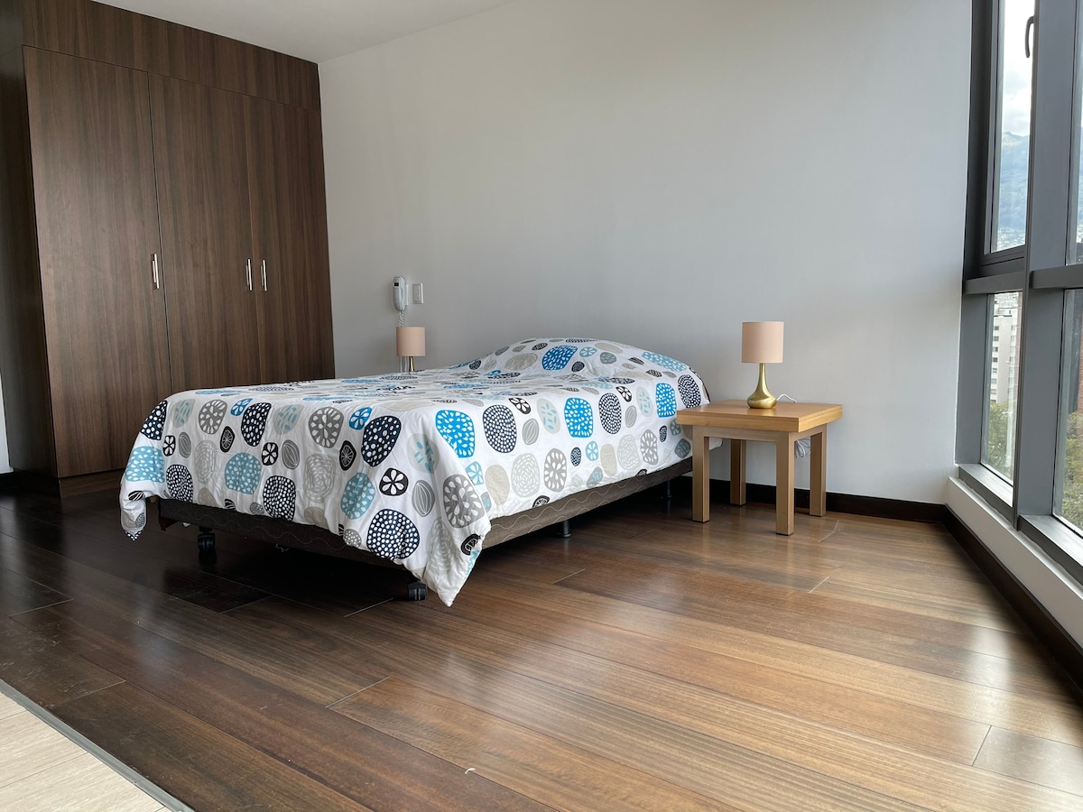 A spacious bedroom features a bed covered with a patterned duvet. A small table with lamps sits beside the bed. Tall windows allow for natural light to illuminate the area, which has wooden flooring and a wardrobe in the background.