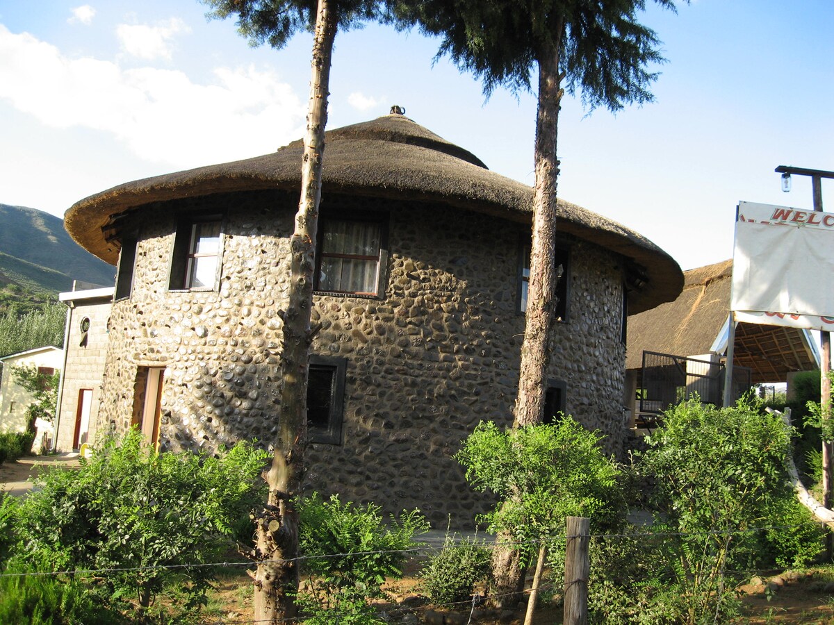 A unique round stone building with a thatched roof is surrounded by lush greenery. Tall trees frame the structure, while windows are evenly spaced along the walls, providing a rustic charm to the exterior.