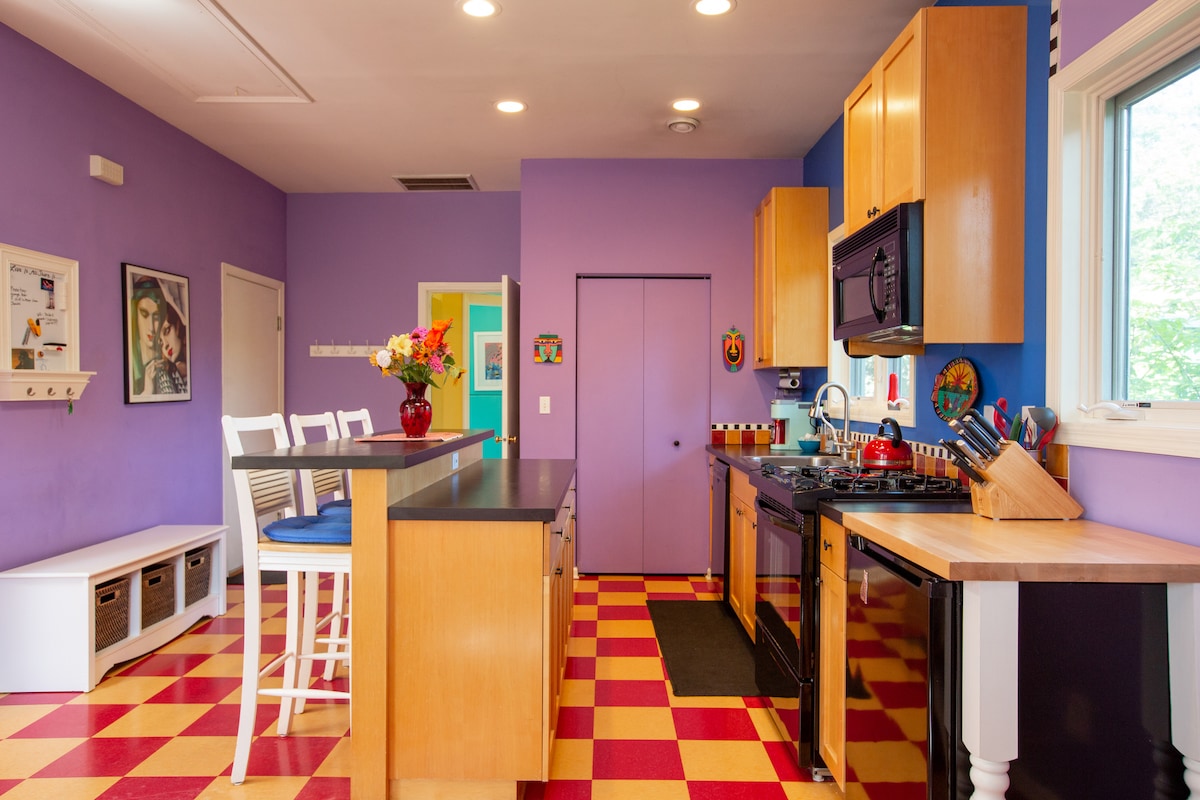 The kitchen features vibrant purple walls complemented by checkered red and yellow flooring. A central island with seating for four is highlighted. The kitchen is fully equipped with black appliances, including a stove and microwave, and ample natural light illuminates the cheerful space through a nearby window.