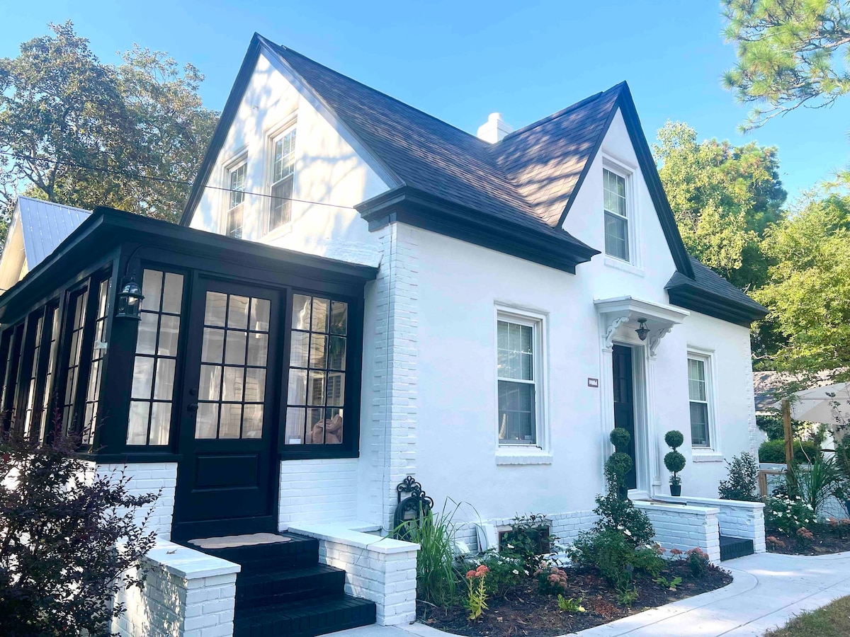 A charming two-story cottage is presented with a white exterior and a black roof. A sunroom with large glass panels is visible at the front, while neatly trimmed shrubs and landscaped gardens enhance the inviting entrance.