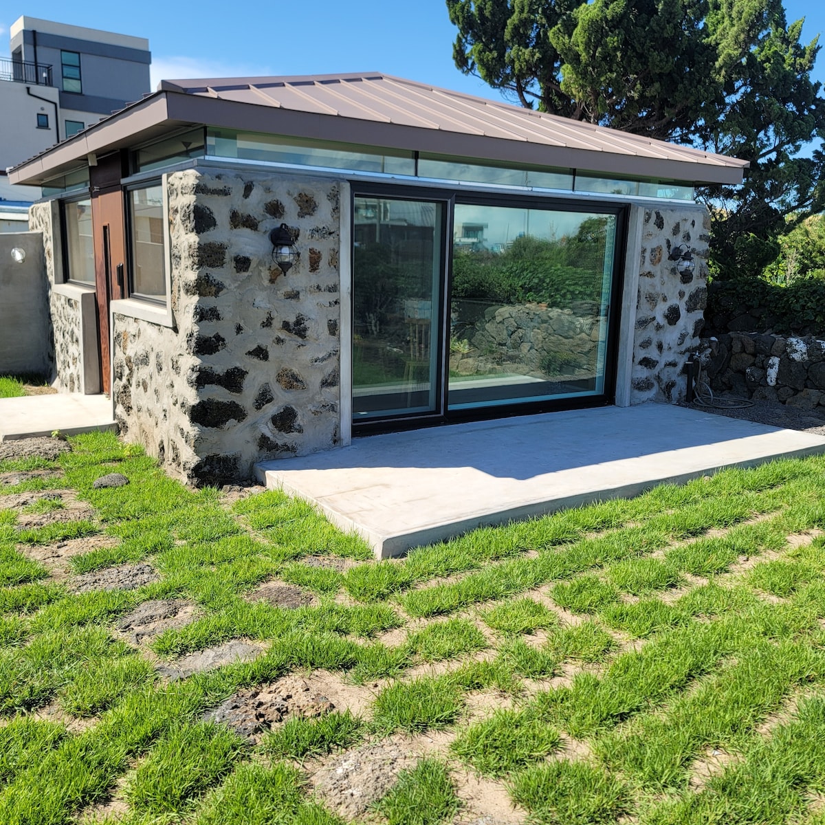 The exterior of a stone-built house is presented, featuring large glass doors that provide a view of the surrounding landscape. The structure is set on a grassy area, with a pathway leading to the entrance. Natural stone walls blend with the modern design.