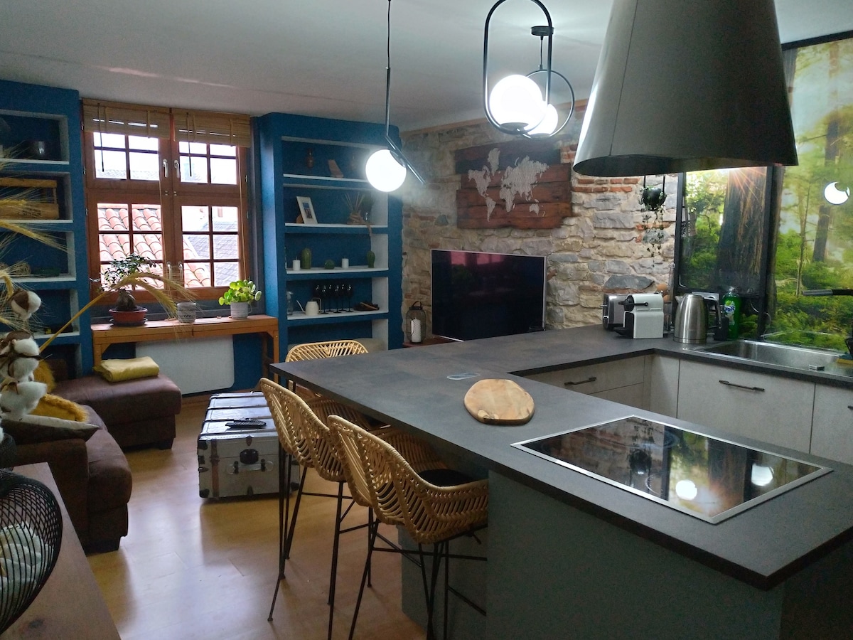 A well-appointed kitchen area showcases modern appliances, including a coffee maker and microwave. A large island with seating is complemented by decorative light fixtures. Natural light filters through the windows, highlighting the warm tones of the stone wall and the greenery from nearby plants.