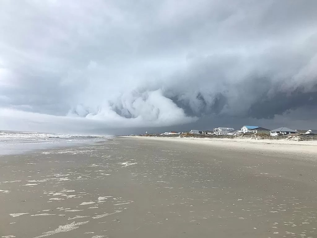 An expansive sandy beach is shown under a dramatic sky filled with thick gray clouds. The shoreline is visible, featuring gentle waves meeting the sand. In the distance, a row of beach houses can be seen, creating a serene coastal scene.