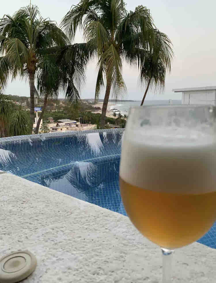 A view of the private jacuzzi is displayed in the foreground, with a glass of beer resting on a textured surface. Palm trees frame the scene, while the ocean stretches out in the background, showcasing the tranquil landscape.