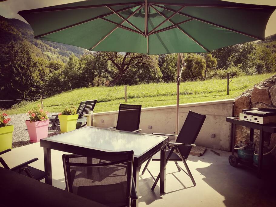 terrasse béton avec plancha gaz et table en verre avec sièges relax, parasol et 2 transats.