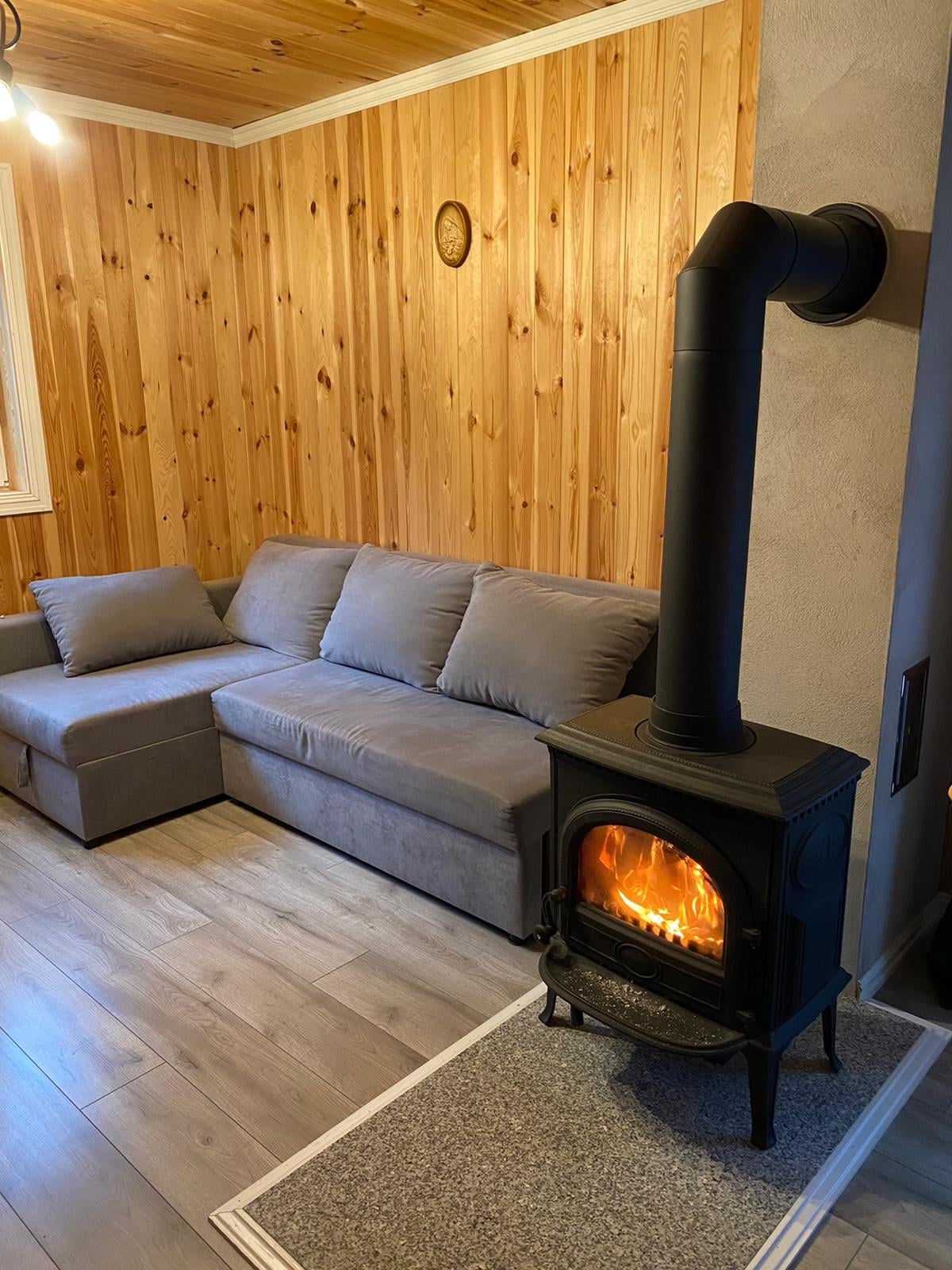 A cozy corner is shown, featuring a gray sofa against a wooden wall. A black stove exudes warmth, with flames visible through its glass front. The wooden ceiling adds a rustic touch to the comfortable ambiance.