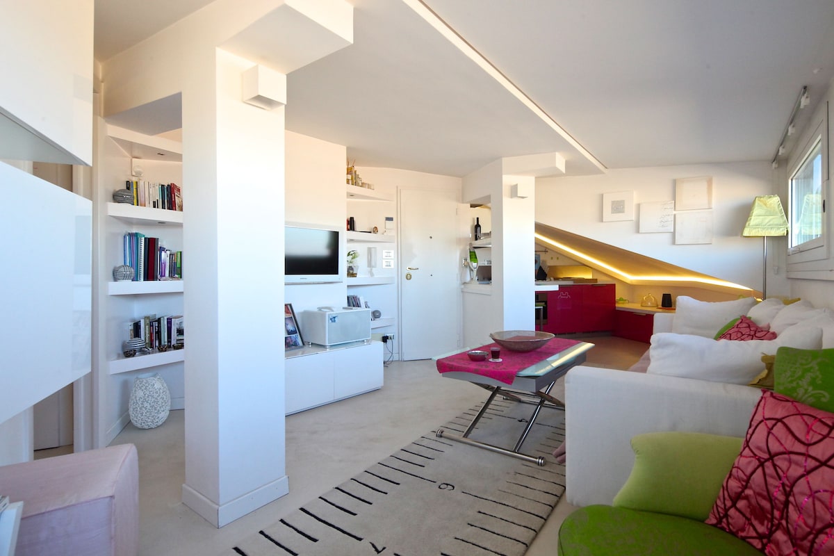 The open living space showcases a comfortable seating area with a white sofa and two armchairs. A vibrant pink table is positioned in the center. Modern shelves display books and decor. Soft lighting highlights the angled ceiling, emphasizing the brightness of the room.